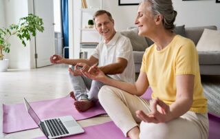 Senior couple sitting on floor in living room meditating during virtual meditation club on laptop