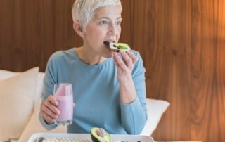 Woman sitting on couch eating avocado toast with a smoothie in the other hand